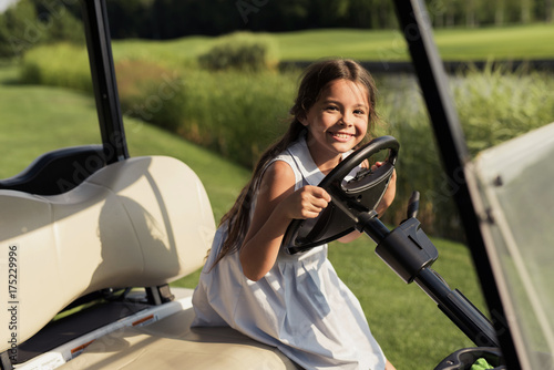 Little girl in a bright dress smiling while sitting at the wheel of a golf car