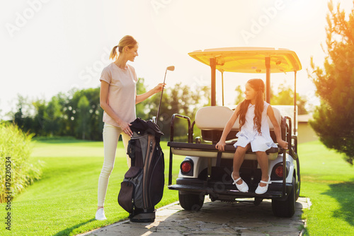 A woman pulls out a golf club from her bag, while the girl sits next to the trunk of a golf cart