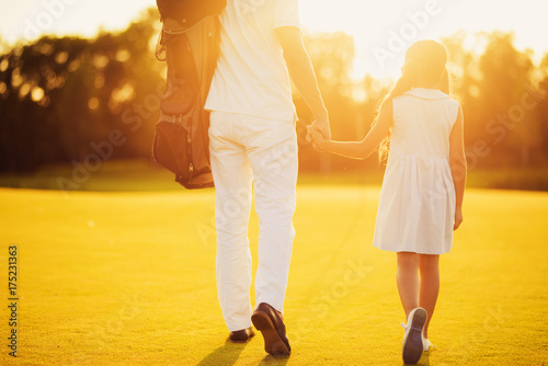 Father and daughter are walking along the golf course against the sunset background holding hands.