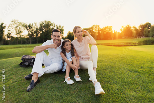 Man, woman and little girl are sitting on the grass on a golf course and smiling