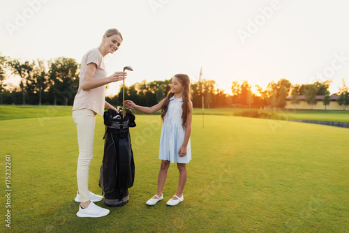 The girl looks at woman who is standing next to her and gives her golf club a bag. They are standing on the golf course
