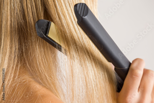 Woman's hand using straightener on her blonde hair on the gray background. Cares about a smooth, beautiful hair. Beauty salon concept.