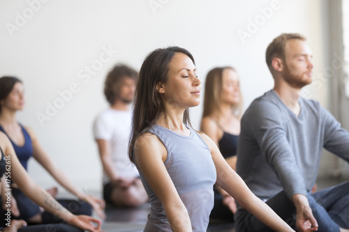 Group of young people, sporty students practicing yoga lesson with instructor, sitting and meditating with closed eyes in Padmasana exercise, Lotus pose, friends working out in club, studio background