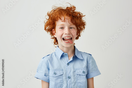 Portrait of silly little ginger boy in blue shirt with wild hair mowing eyes, smiling and showing tongue in camera,making funny faces.