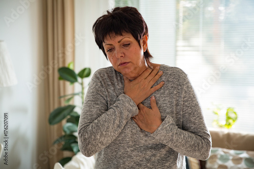 Sick mature woman with sore throat, standing in living room at home. Catching cold, having cough.