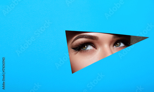 A girl with beautiful green bright eyes with brown shadows and expressive eyebrows looks into the hole of colored paper.Fashion, beauty, make-up, cosmetics, make-up artist, beauty salon,business card.