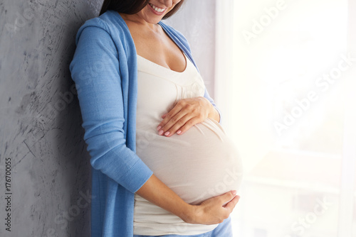 Smiling pregnant woman holding belly while posing against grey background