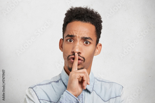 Serious dark skinned male keeps finger on lips, asks to be silent and not interrupt him, isolated over white background. Fashionable attractive young man makes hush sign, has confident expression