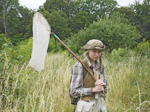 A girl entomologist in a field near a forest. A young woman is dressed in country style, has two plaits, wears a hat and holds an insect net. A student summer field period in the university.