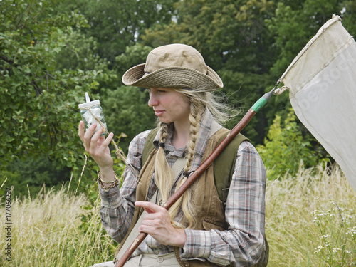 A girl entomologist in a field near a forest. Young woman with two plaits, in a hat, dressed in country style holds an insect net and a killing bottle. A student summer field period in the university.