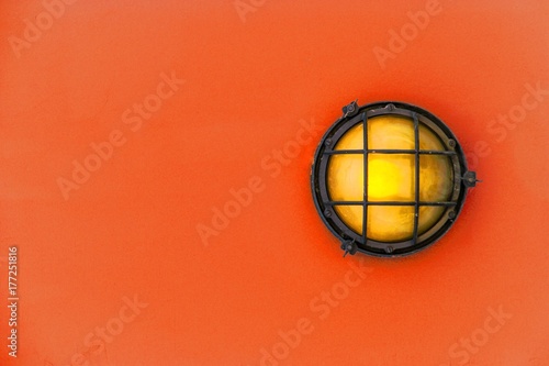 Yellow bulkhead light (ship deck lamp) surrounded by a metal rusted frame fixed to a painted red orange color wooden wall background.