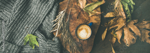 Autumn or Fall morning coffee concept. Flat-lay of knitted woolen grey sweater, wooden tray, mug of coffee and yellow fallen leaves over dark rustic wooden table background, top view, wide composition