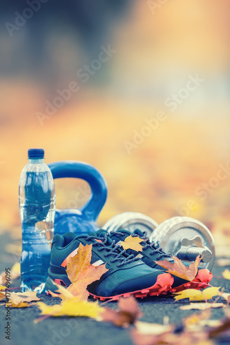 Pair of blue sport shoes water and dumbbells laid on a path in a tree autumn alley with maple leaves - accessories for run exercise or workout activity