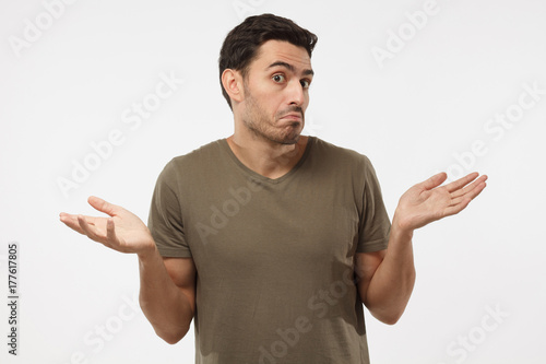 I don't know. Young man isolated on grey background being at a loss, showing helpless gesture with arm and hands, mouth curved as if he does not know what to do