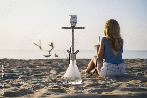 A woman smoking shisha (hookah) sitting by the sea. Seagulls at the background.