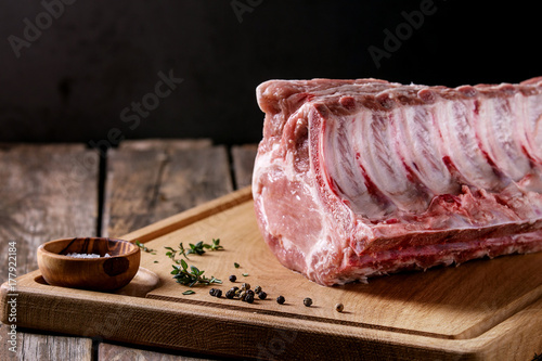 Fresh raw uncooked whole rack of pork loin with ribs on wooden cutting board with salt, thyme and butcher clever over old wood plank table. Close up.