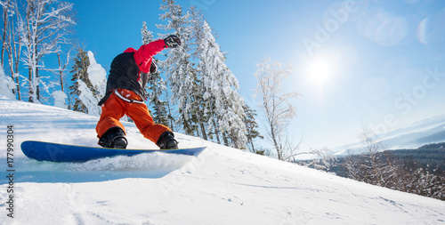 Horizontal low angle shot of a male snowboarder riding the slope on a sunny winter day in the mountains. Forest, blue sky and sun on the background. copyspace freeride snowboarding active lifestyle