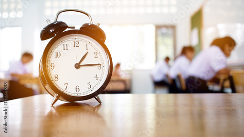 Retro alarm clock with one o'clock, left side on wood table teacher in blur students exams classroom. Time is indefinite continued progress of existence. Education exams concept, black and white