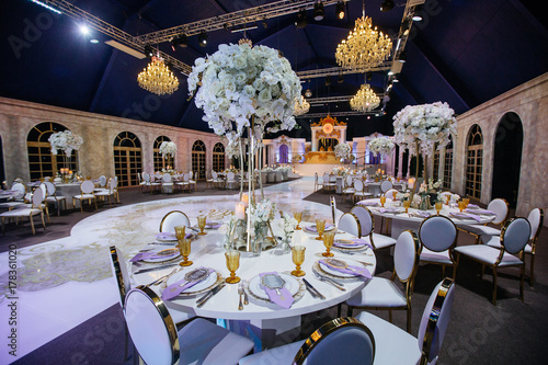 Large white bouquets of roses stand on dinner tables decorated with golden glasses and violet cloth