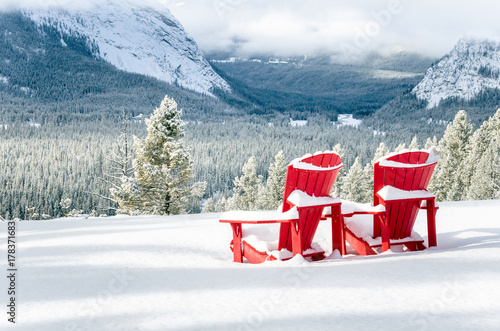 Snow Covered Red Adirondack Chairs Facing a Frozen Forested Valley on a Snowy Winter Day. Banff National Park, AB, Canada.