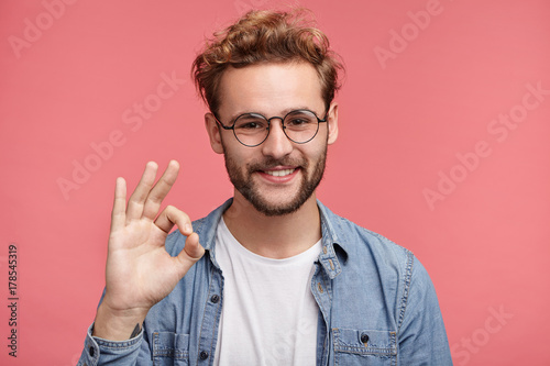 Positive hipster guy shows ok sign, demonstares that everything is fine, agrees with people who surrounds him. Confident cheerful man gestures indoors. Body language and human emotions concept