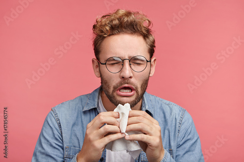 Portrait of ill or sick bearded male sneezes and coughs, uses handkerchief, rubs nose, being allergic to animals. Man has running nose, caught cold when had walk outdoors in winter or autumn