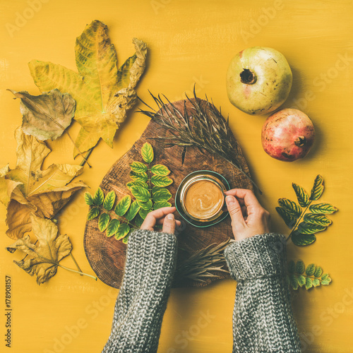 Autumn morning coffee concept. Flat-lay of woman' s hands in grey woolen sweater holding cup of espresso over mustard yellow background with dried leaves and pomegranates around, top view, square crop