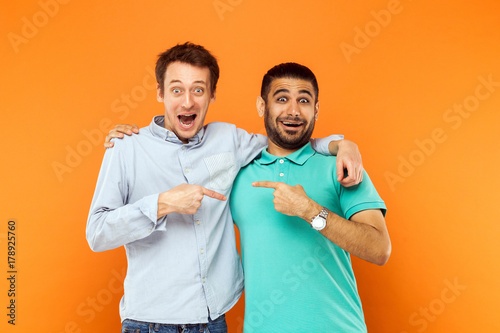 Two best friends hugging, pointing finger each other and looking at camera with shocked face