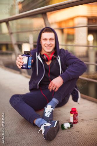 The young sporty man sitting on the floor outdoor and holding protein pills in hand