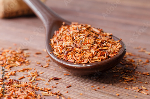 Composition of Rooibos tea is located on a ceramic spoon. Macro photo of a tea close-up. The petals of tea are located in a spoon on a wooden board. Natural ingredients of Rooibos tea.