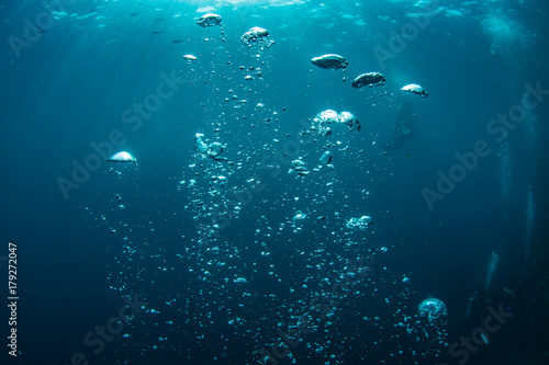 Underwater shot of blue ocean water, air bubbles closeup, sunbeams on water surface, some scubadivers in deep
