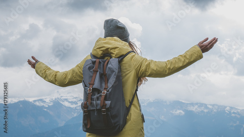 Young Caucasian female hiker in yellow raincoat wearing backpack enjoys the mountain view in French Alps