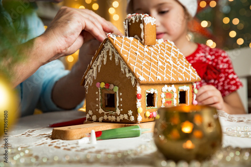 Father and adorable daughter in red hat building gingerbread house together. Beautiful decorated room with lights and Christmas tree, table with candles and lanterns. Happy family celebrating holiday.