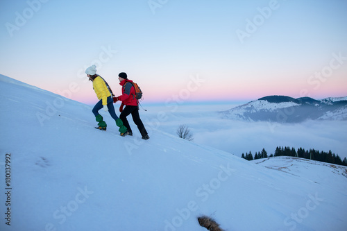 Young couple hiking outdoors with backpacks in winter mountains. Rear view shot of young woman with her boyfriend in the mountains. Hikers with backpacks enjoying sunset. Travel concept