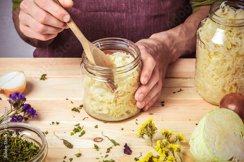 The cook puts sauerkraut in glass jars. On the table are spices, onions and cabbage.