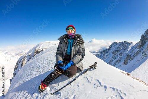 woman climber in helmet and down jacket with trekking sticks sits on top of a mountain