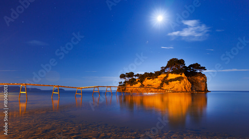 Small tropical island with wooden hanging bridge from Zante Greek island to it. Panoramic view of Night scenery beneath the moon. Aegean sea. Zante Zakinthos is popular and famous Greek resort.