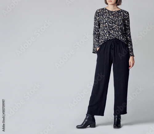 Woman wearing stylish outfit with black patterned blouse, black high-waisted wide leg trousers and black ankle boots isolated on grey background. Copy space