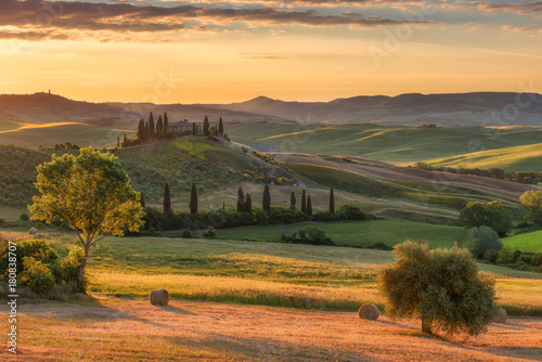 Magnificent spring landscape at sunrise.Beautiful view of typical tuscan farm house, green wave hills, cypresses trees, hay bales, olive trees, beautiful golden fields and meadows.Italy, Europe