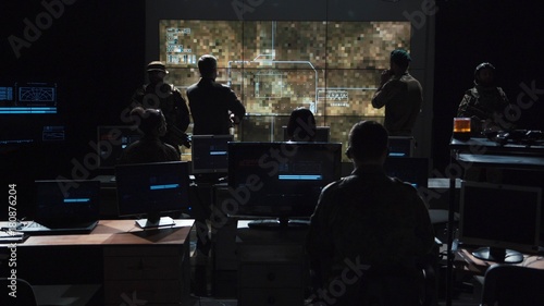 Group of soldiers or spies in dark room with large monitors and advanced satellite communication technology launching a missle. Includes flashing yellow light.