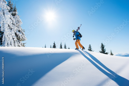 Low angle shot of a skier walking on top of the mountain with his skis on his shoulder on a sunny winter day sunlight nature recreation skiing lifestyle downhill sportive concept