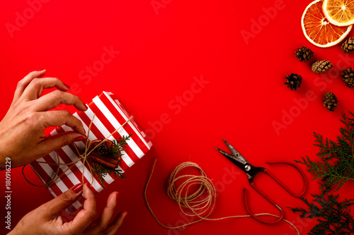 Female Hands Holding Festive Christmas Box Twine Cord on the Red Paper Background Holiday Top View Copy space for Text Advent Gift Flat Lay