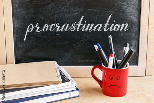Lettering of word Procrastination on black chalkboard on a background of notepads and diary and a red cup with a sad smile. Business and pischological concept. Self-control and achieving the goal