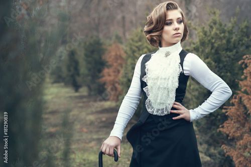 Portrait of gorgeous young woman with elegant Victorian hairstyle wearing old-fashioned gown with jabot leaning on her cane and looking aside, misty autumn park on background. Vintage style