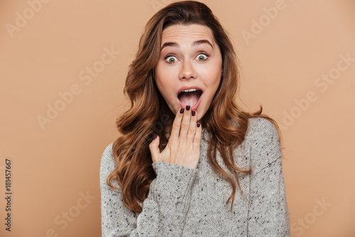 Close-up photo of amazed pretty girl in gray knitted sweater covering her mouth while looking at camera