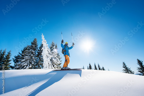 Low angle shot of a cheerful skier holding ski poles raising his arms in the air achievement leadership celebration winning victorious happiness positivity gesturing resort riding lifestyle extreme