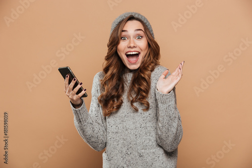 Portrait of happy exited brunette girl in gray hat holding mobile phone while looking at camera