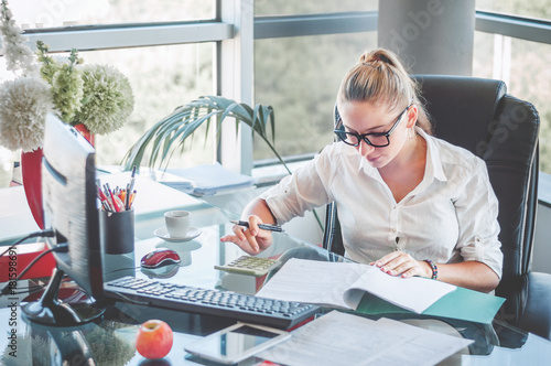 Portrait of young business lady in glasses sitting at her workplace and reads documents. Office worker at modern office workplace. Business concept.