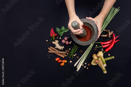 The Art of Thai Cuisine - Thai lady’s hands hold stone granite pestle with mortar and red curry paste ingredient together with fresh herbs and spices on classic dark background at top view angle.