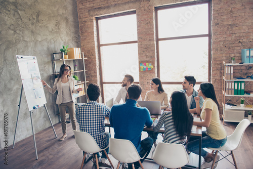 Young smart lady in glasses is reporting to the team of colleagues about the new project at the meeting with the white board. Workers are listening to her, all dressed in casual outfits